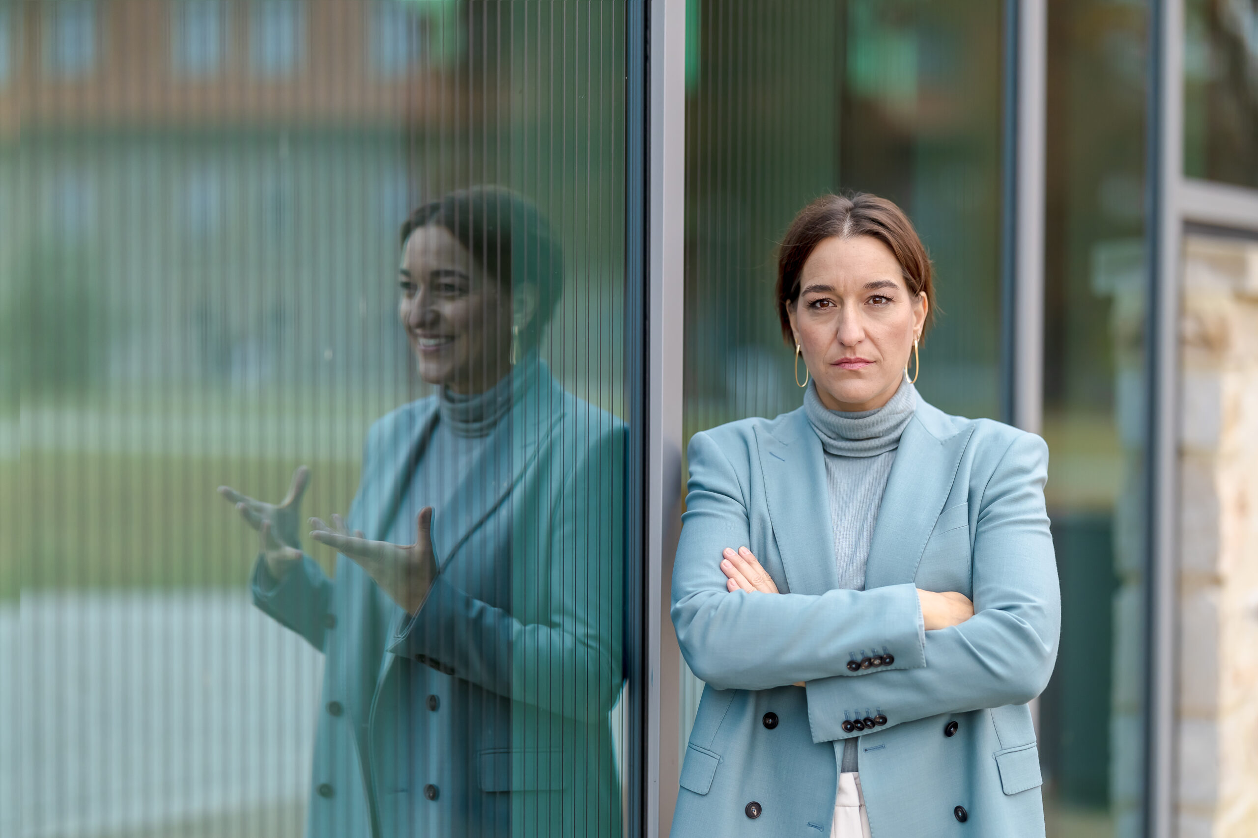 Katinka Riemann mit verschränkten Armen und ernstem Blick vor einer Glasfassade, ihr Spiegelbild gestikuliert