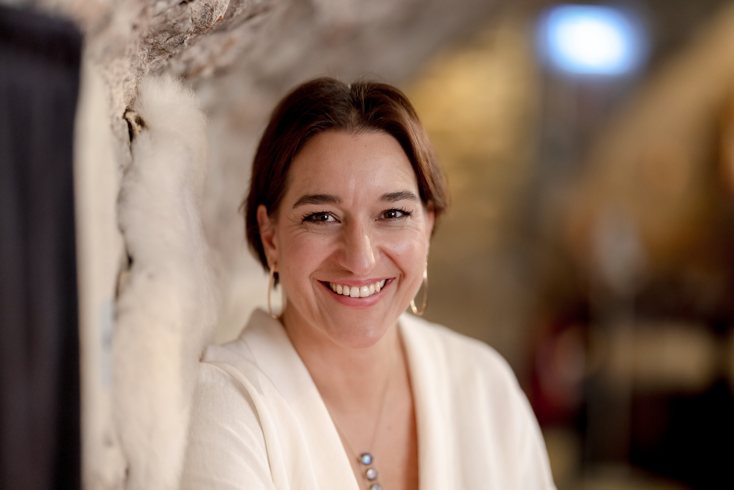 Porträt von Katinka Riemann in heller Strickjacke, lächelnd an eine Steinwand gelehnt