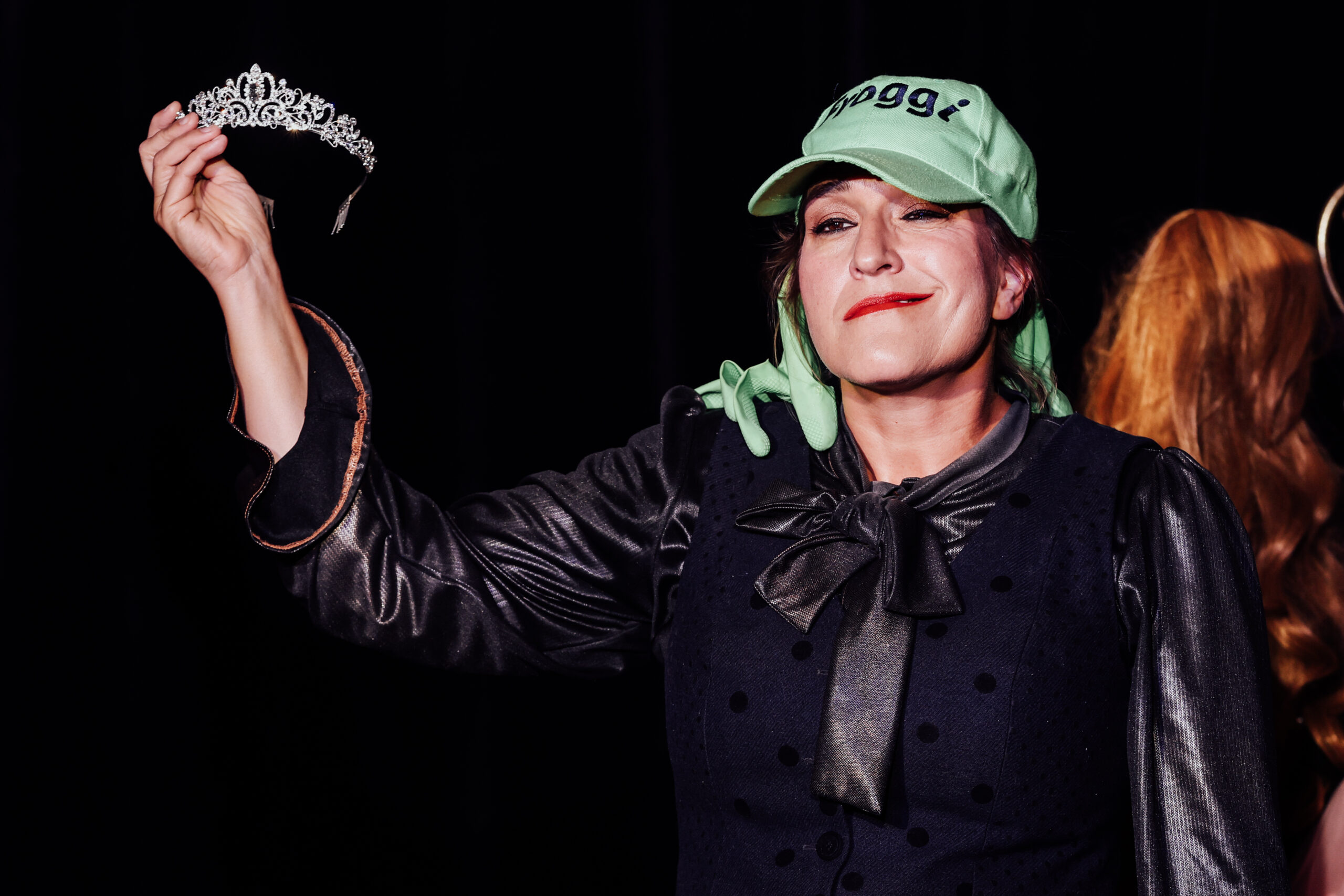 Katinka Riemann in „Froschsalat“ mit grüner Schirmmütze, hält lächelnd ein Diadem hoch