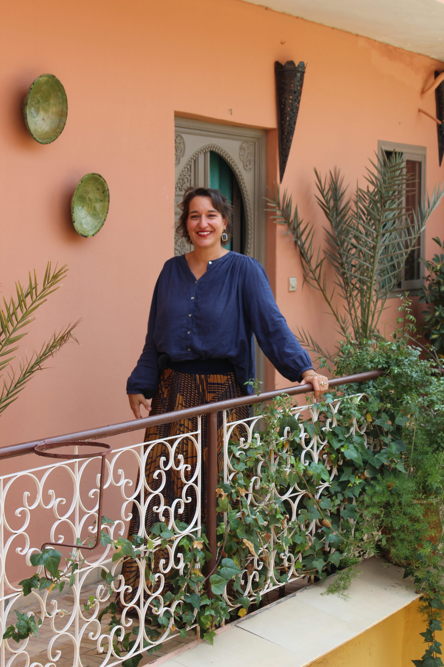 Retreat-Leiterin in blauer Bluse auf einem Riad-Balkon in Marrakesch, hinter ihr eine terrakottafarbene Wand