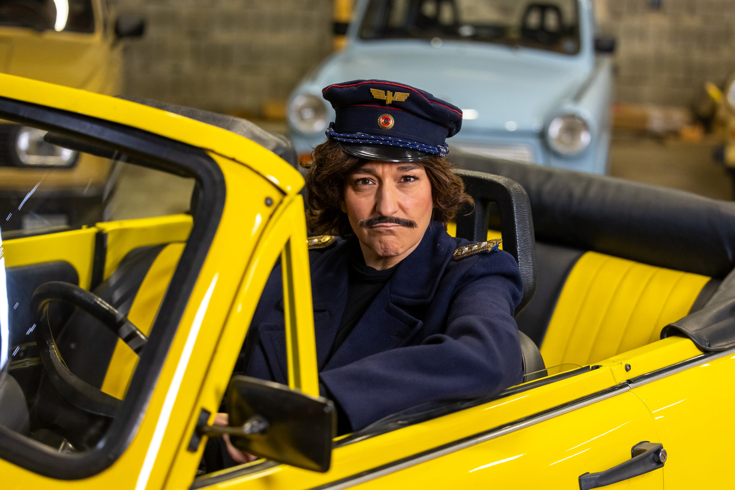Katinka Riemann in einer Rolle mit Schnurrbart und Uniformmütze im gelben Cabrio